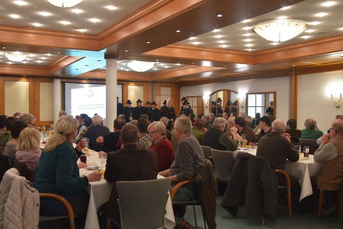 Eine große Gruppe von Menschen sitzt an Tischen in einem Saal während einer Jahresversammlung. An der Wand hängt ein Bildschirm mit dem Titel „Jahreshauptversammlung Hegegemeinschaft Bopfingen 2018“. Im Hintergrund stehen einige Personen in schwarz gekleideten Uniformen.